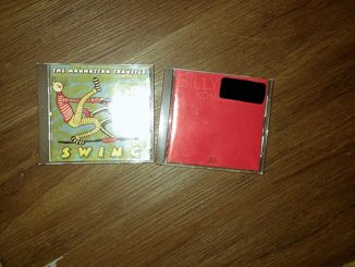 Two cd albums on a brown floor, on the left is Swing by The Manhattan Transfer, and on the right is Концерт (Kontsert) by Billy Joel. Swing's album art is of two loopy cartoon figures dancing together with The Manhattan Transfer written on the top, and Swing written on the bottom, both in whimsically unserious fonts. Camera flash obscures the right third of the art, but barely misses any text. Концерт's album art is a pure red, the title of the album and Billy Joel's name is written on it in a very similar red, making it hard to read. There is a black rectangle on the upper right corner of Концерт as a censor bar covering a barcode and store label.